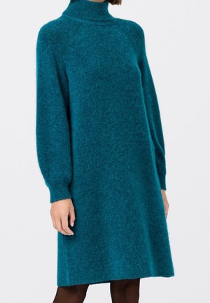 Kobieta ubrana w miękki, długi turkusowy sweter z golfem, stojąca na jednolitym białym tle.