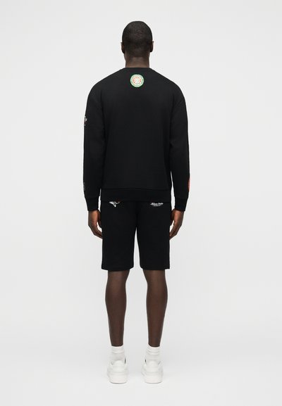 Άνδρας που στέκεται με την πλάτη προς τα εμπρός, φοράει μαύρη μπλούζα μακρυμάνικη με κυκλικό λογότυπο στο πάνω μέρος της πλάτης, μαύρα σορτς, λευκές κάλτσες και λευκά αθλητικά παπούτσια.