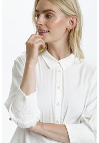 Chemise blanche à bouton avec col classique et poignets élastiqués, présentant une texture lisse et une coupe décontractée.