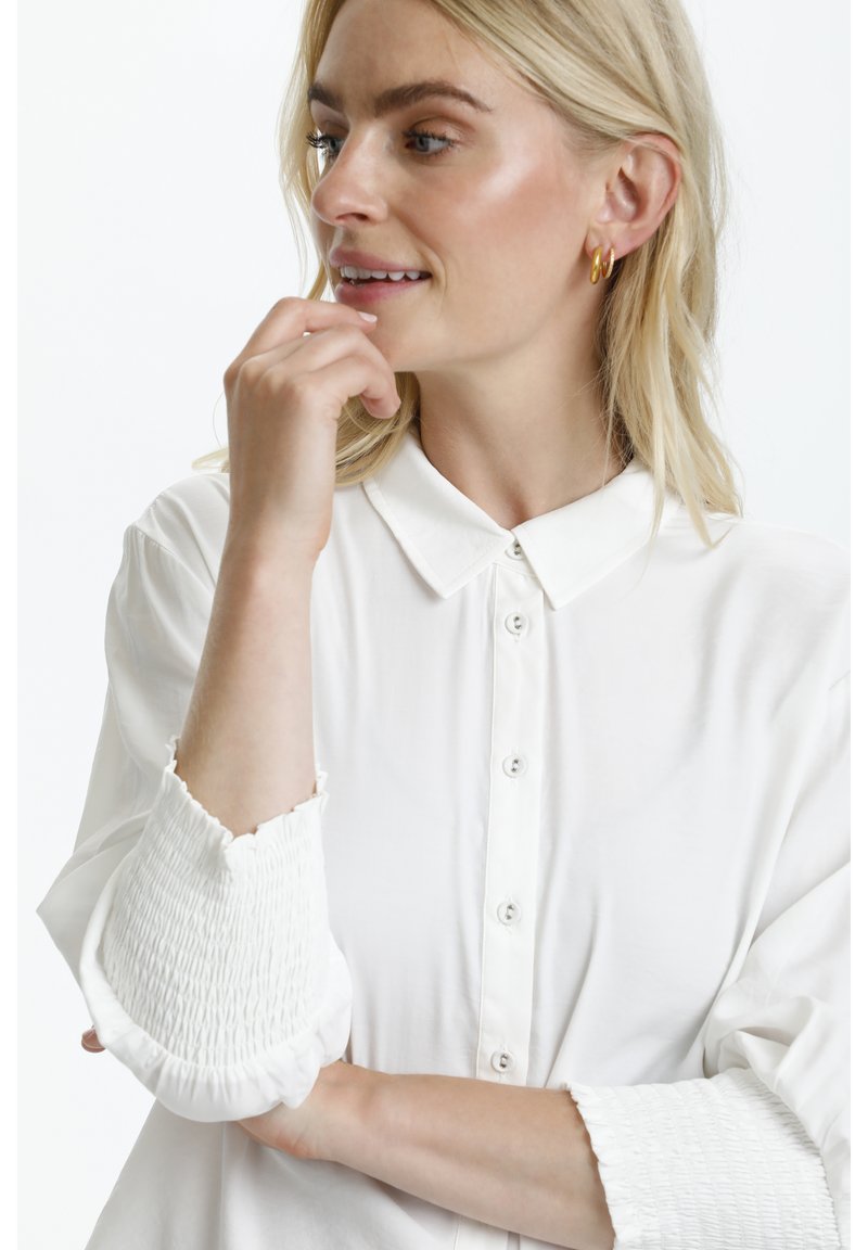 Chemise blanche à bouton avec col classique et poignets élastiqués, présentant une texture lisse et une coupe décontractée.