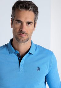 Hellblaues Poloshirt aus glattem Baumwollmaterial mit einem dunkelblauen Kragen, versehen mit einem kleinen gestickten Logo auf der linken Brust.