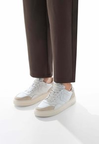 Sneakers bianche in pelle con accenti in suede beige, punta perforata, lacci piatti e suole in gomma texture, abbinate a pantaloni marroni.