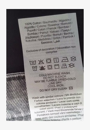 Bijela oznaka za njegu s crnim tekstom koji navodi upute za njegu na više jezika. Sadrži simbole za pranje, sušenje i peglanje. 100% pamuk.