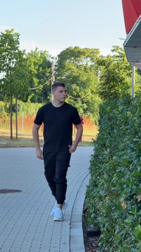 Melnā T-kreklā, melnās cargo biksēs un baltos sporta apavos. Cilvēks staigā pa bruģētu taku blakus zaļai dzīvžogam un kokiem.