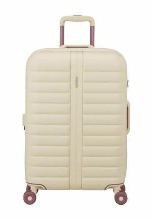 Valise rigide couleur crème avec poignée télescopique violette et quatre roues pivotantes violettes, debout sur fond blanc.