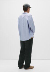 Lyseblå og hvide stribede oversized skjorte, sorte jeans og beige sko. Grå kasket, set fra ryggen mod en plain hvid baggrund.