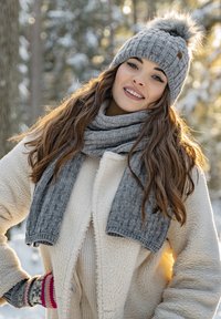 Bonnet en tricot gris avec un pompon, écharpe grise assortie avec une texture côtelée, et manteau en polaire crème dans un décor de forêt enneigée.