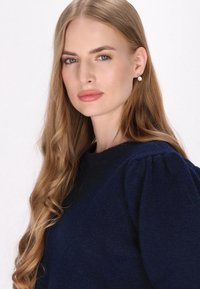 Tengerés kék kötött pulóver kerek nyakkivágással és puffos ujjakkal. A modellen hosszú, hullámos haj található, és gyöngy fülbevalót visel.