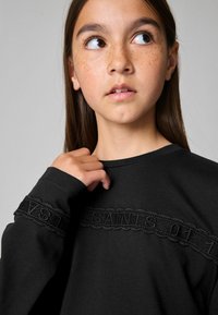 Must have pikkade varrukatega must pluus tekstuuriga kangast ja dekoratiivse musta pitsiriba, millel on rinnaosas sõna "SAINTS".