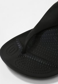 Tongs noires avec une bride lisse, semelle texturée avec des points et motifs en relief, et un design plat pour un confort et un chaussant faciles.