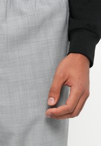 Tissu gris avec un motif de tissage subtil, positionné à la main près de la taille, portant une manche à poignets noire.