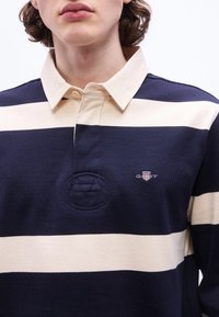 Νεαρό άτομο που φορά μπλούζα ράγκμπι με ναυτικό μπλε και μπεζ ρίγες και μπεζ γιακά, με κεντημένο λογότυπο στο στήθος.