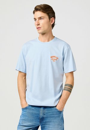 Helles blaues Baumwoll-T-Shirt mit Rundhalsausschnitt und kurzen Ärmeln. Verfügt über ein oranges Logo "Wrangler Jeans 1947" auf der Brust.