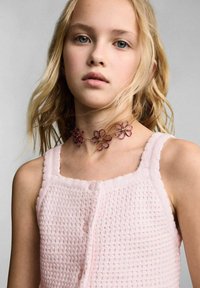 Roza pletena majica z teksturiranim vzorcem in gumbnim zapenjanjem. Nosila je cvetlični ovratnik z tremi vijoličnimi kovinskimi cvetovi.