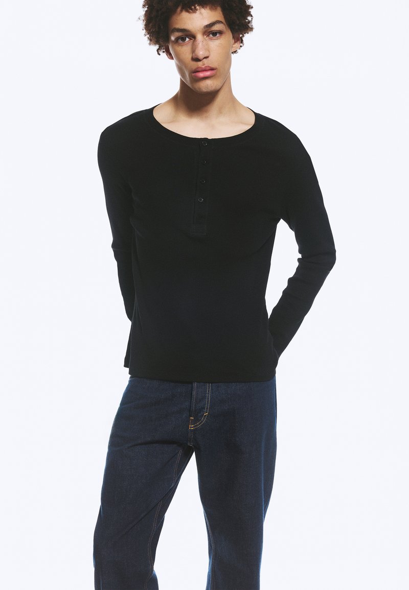 Jeune homme aux cheveux bouclés portant un t-shirt henley noir à manches longues et un jean bleu foncé, debout les mains dans le dos.