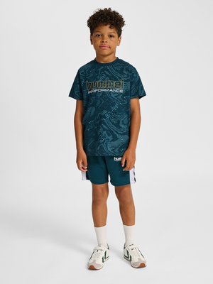 Dreng stående med krøllet hår, iført en mørkeblå mønstret Hummel Performance T-shirt, matchende shorts, hvide sokker og hvide sneakers.