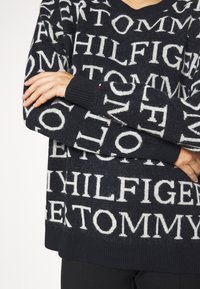 Marinefarbener Pullover mit großem weißen Schriftzug "TOMMY HILFIGER". V-Ausschnitt und gerippte Bündchen, aus weichem Stoff mit leichter Textur.