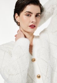 Biały, puszysty kardigan w romby, z złotymi guzikami i teksturowanym materiałem. Modelka pozuje z ręką na szyi.