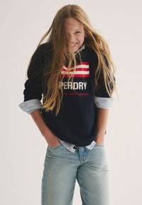 Maglione a maglia navy con strisce rosse e bianche e logo "Superdry". Abbinato a jeans azzurri e una camicia a righe blu sotto.
