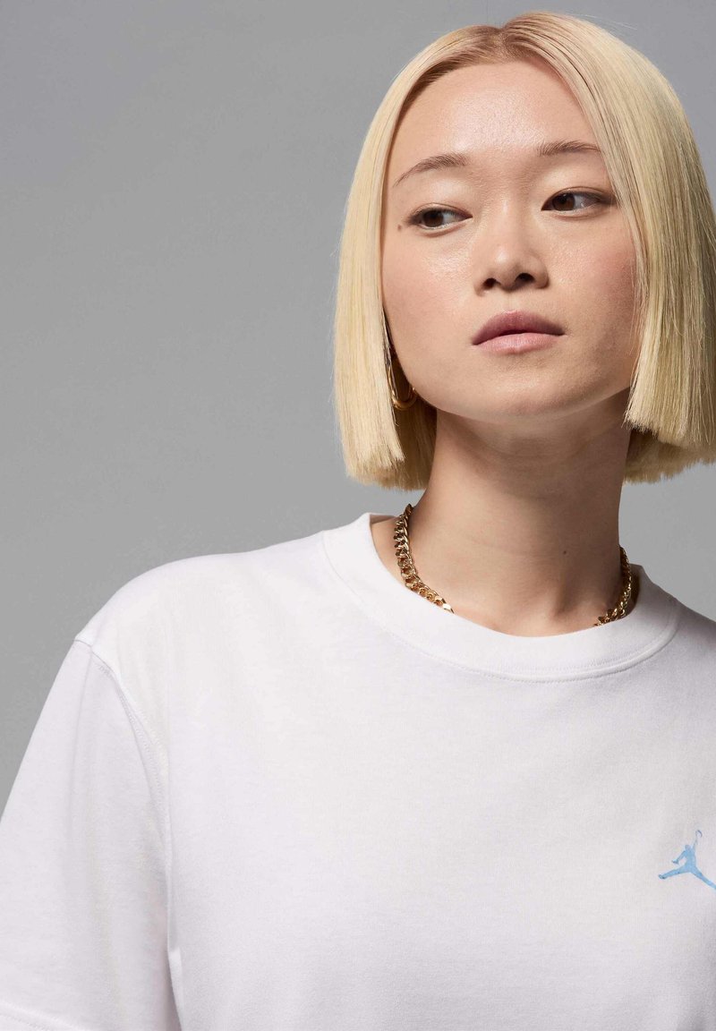 Camiseta de algodón blanca de manga corta, con un logo azul claro en la parte izquierda del pecho; llevada con un collar de cadena dorada.