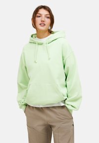 Felpa verde chiaro con cappuccio e lacci, tasca frontale e logo minimale. Indossata sopra una maglietta bianca, abbinata a pantaloni cargo beige.