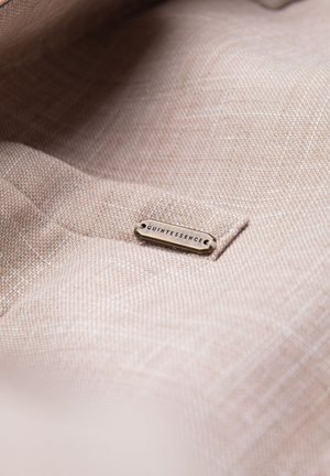 Lichtbruine textuurstof met een klein metalen label erop genaaid, gegraveerd met het woord "QUINTESSENCE."