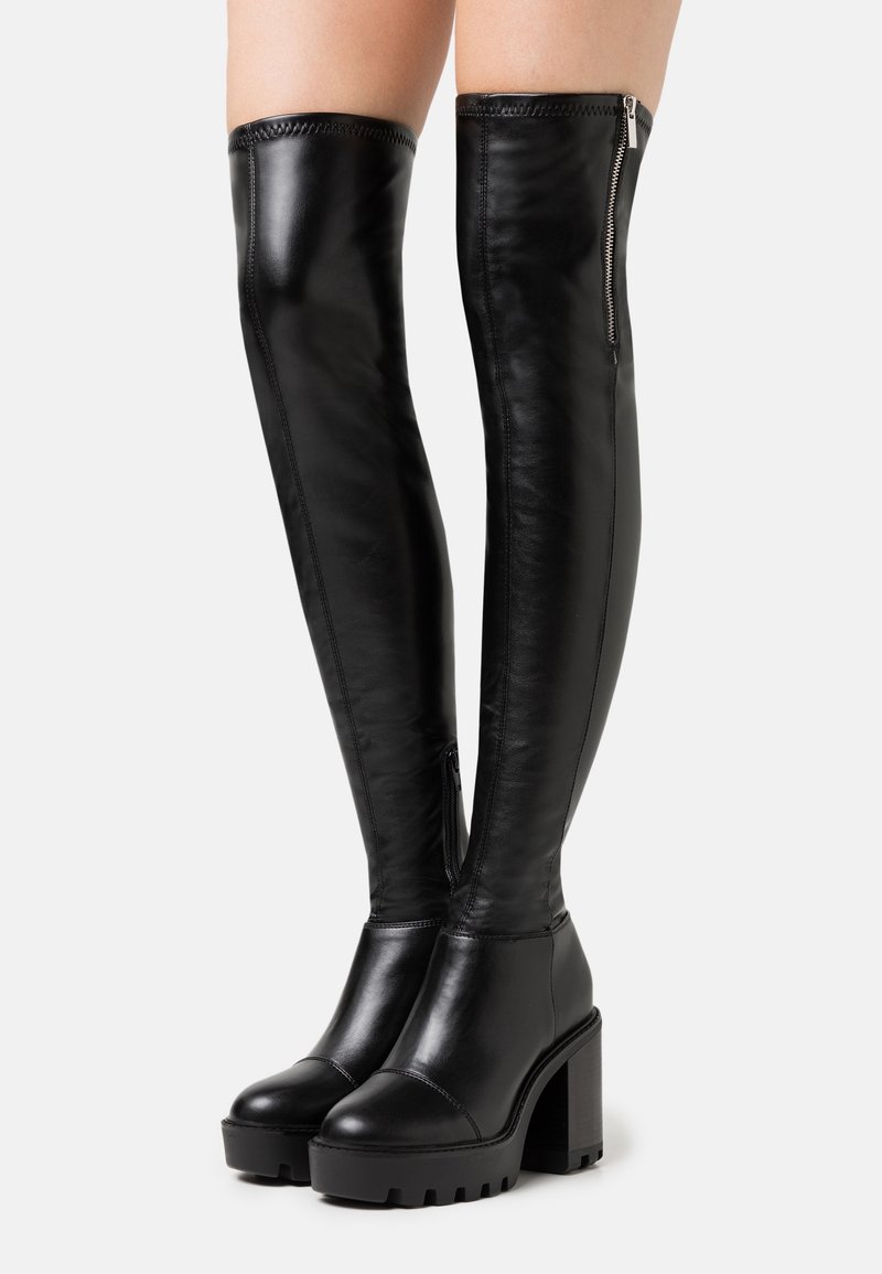 Bottes noires en cuir montantes au-dessus du genou avec plateforme, talons hauts épais et fermeture éclair latérale, portées sur des jambes nues sur un fond clair.