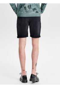 Persona che indossa pantaloni corti neri arrotolati, scarpe nere stringate e un maglione verde con stampe botaniche, con tatuaggi visibili sulla parte posteriore dei polpacci.