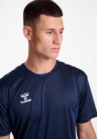 Marineblå sports trøje med korte ærmer, V-hals design og kontrasterende blå detaljer. Har Hummel-logoet på venstre bryst.