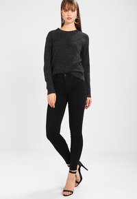 Mustat slim-fit farkut yhdistettynä tummanharmaaseen, teksturoituun neuleeseen. Mallilla on mustat korkeat avokkaat ja renkaanjuhlat korvissa, ja hän seisoo yksinkertaista taustaa vasten.