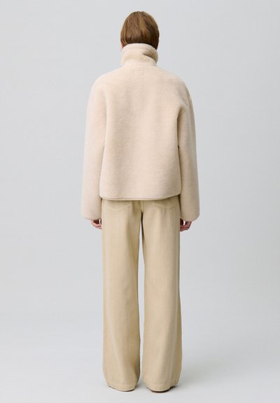 Giacca in pile beige con colletto alto, vestibilità rilassata e superficie testurizzata. Abbinata a pantaloni in corduroy a gamba larga di colore crema.