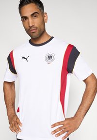 Λευκό αθλητικό πουκάμισο με μαύρα, κόκκινα και κίτρινα πάνελ ως διακοσμητικά στοιχεία, με στρογγυλή λαιμόκοψη και ένα εξέχον έμβλημα της γερμανικής ποδοσφαιρικής ομάδας στο στήθος.