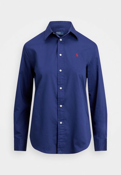 Tumši zils garām piedurknēm krekls, kas izgatavots no kokvilnas, ar pogājamu priekšpusi, norādītu apkakli un mazu sarkanu logotipu kreisajā krūtī.