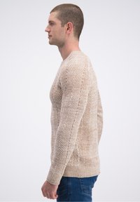 Beiger Pullover mit Fischgrätmuster, Rundhalsausschnitt und gerippten Bündchen. Gesehen von der Seite, getragen mit blauer Jeans.