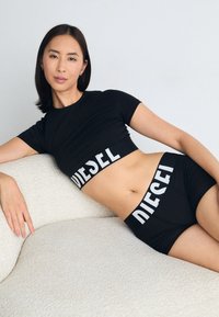 Juodas trumpas marškinėlis ir šortai su balta "DIESEL" logotipu ant juosmens. Lygus, prigludęs audinys; modernus ir sportiškas dizainas.