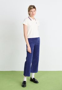 Polo en crème avec un col bicolore ; assorti à un pantalon en velours côtelé bleu marine à jambe large ; complété par des mocassins noirs et des chaussettes blanches.