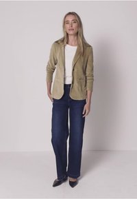 Chaqueta de pana beige sobre una camisa blanca, combinada con jeans de pierna ancha azul oscuro y zapatos negros puntiagudos. Textura suave y diseño entallado.