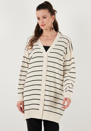 Cárdigan de canalé crema con rayas negras, cuello en V, mangas largas y grandes botones en la parte delantera. La tela tiene una textura suave y un corte relajado.