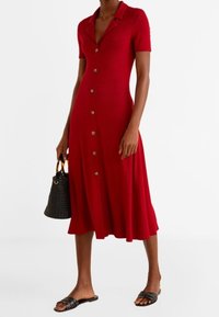 Rotes Hemdblusenkleid mit Kragen, kurzen Ärmeln, ausgestellt geschnittenem Saum und goldenen Knöpfen. Kombiniert mit einer schwarzen Lochmuster-Tasche und flachen Sandalen.