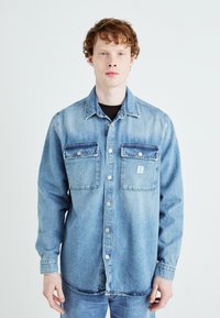 Camicia di jeans azzurro chiaro con due tasche sul petto, bottoni a pressione e una texture leggermente sbiadita. Maniche lunghe con polsini bottonati.