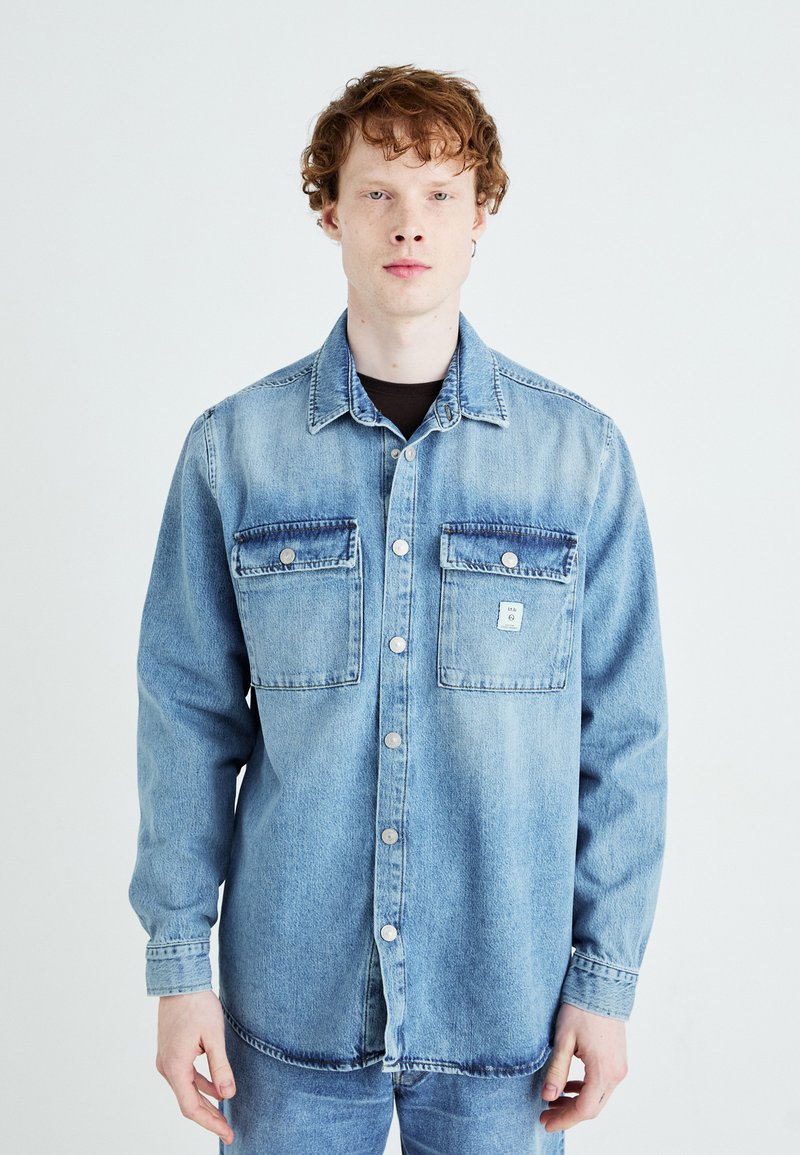 Camicia di jeans azzurro chiaro con due tasche sul petto, bottoni a pressione e una texture leggermente sbiadita. Maniche lunghe con polsini bottonati.