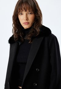 Cappotto doppiopetto nero con un morbido colletto in pelliccia sintetica nera, design strutturato e texture liscia, abbinato a un dolcevita nero a coste.