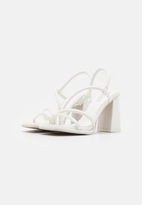 Sandales à talons blancs avec plusieurs fines lanières, un talon bloc et une finition en cuir lisse. La semelle est plate avec un design minimaliste.