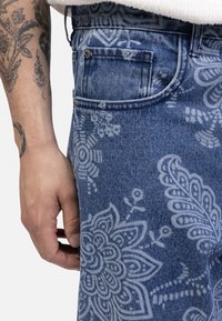 Denim jeans with a light blue base, featuring white floral and paisley patterns. Includes a side pocket and visible stitching detail.