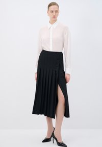 Gonna nera a pieghe con uno spacco laterale e dettagli con bottoni, abbinata a una camicia bianca trasparente e scarpe con tacco nere.