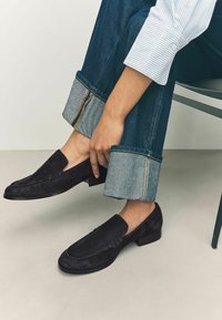 Mocassini in suede neri con tacco basso, texture liscia e dettagli cuciti, abbinati a jeans in denim con orli ripiegati e cuciture a contrasto.