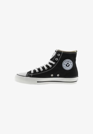 Zapatilla de lona negra de caña alta con suela de goma blanca, puntera redonda, cordones negros y parche con el logo que presenta el texto "Victoria" en el lateral.