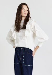 Blusa bianca con dettagli arricciati, rifiniture in pizzo intricate e maniche strutturate, abbinata a jeans blu scuro con borchie decorative multicolori.