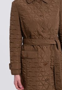 Abrigo exterior acolchado marrón con cierre de botones, bolsillos laterales, cuello y un cinturón a juego. Presenta una textura de patrón de diamantes.