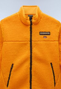 Bright orange fleece jacket with a high collar, two zippered side pockets, and a contrasting dark zipper. Features brand logo and flag detail.